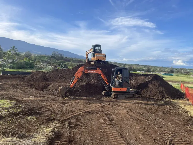 Excavator working a large soil pile on a cleared Maui lot with the valley behind