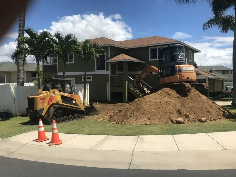 Skid steer and excavator moving a soil pile in front of a finished two-storey home on Maui