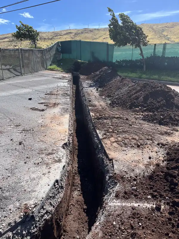 Narrow, deep utility trench cut along the edge of a paved road with the West Maui mountains behind